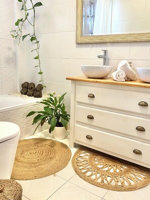 A bathroom with a white sink, two bowls, a rolled towel, a potted plant, and a mirror.