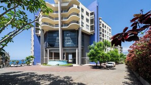 Ein modernes Gebäude mit geschwungener Fassade, großen Fenstern und einer gepflasterten Fläche mit Bäumen und einem kleinen Pool.