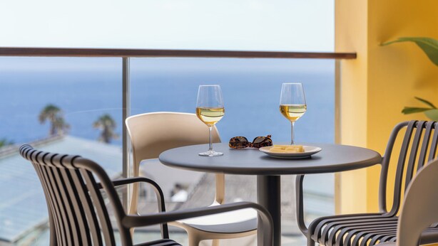 Ein Balkon mit zwei Stühlen, einem Tisch, zwei Weingläsern und einer Käseplatte.