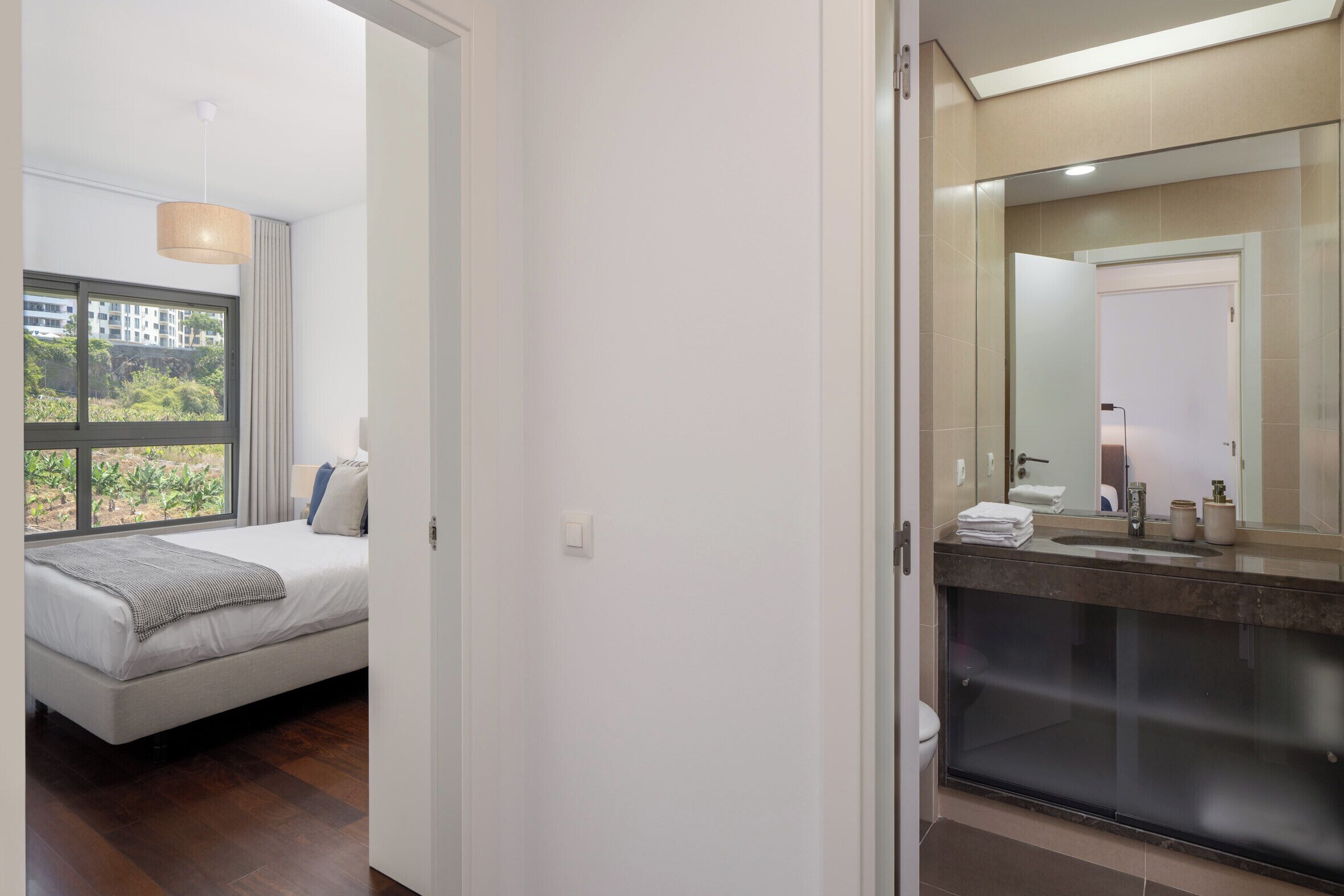 A modern hotel room with a large bed, a bathroom with a glass shower enclosure, and a view of greenery through the window.