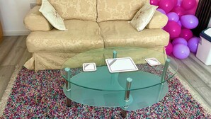 Un divano beige con motivi floreali e due cuscini, un tavolino in vetro con carte da gioco e un tappeto colorato.