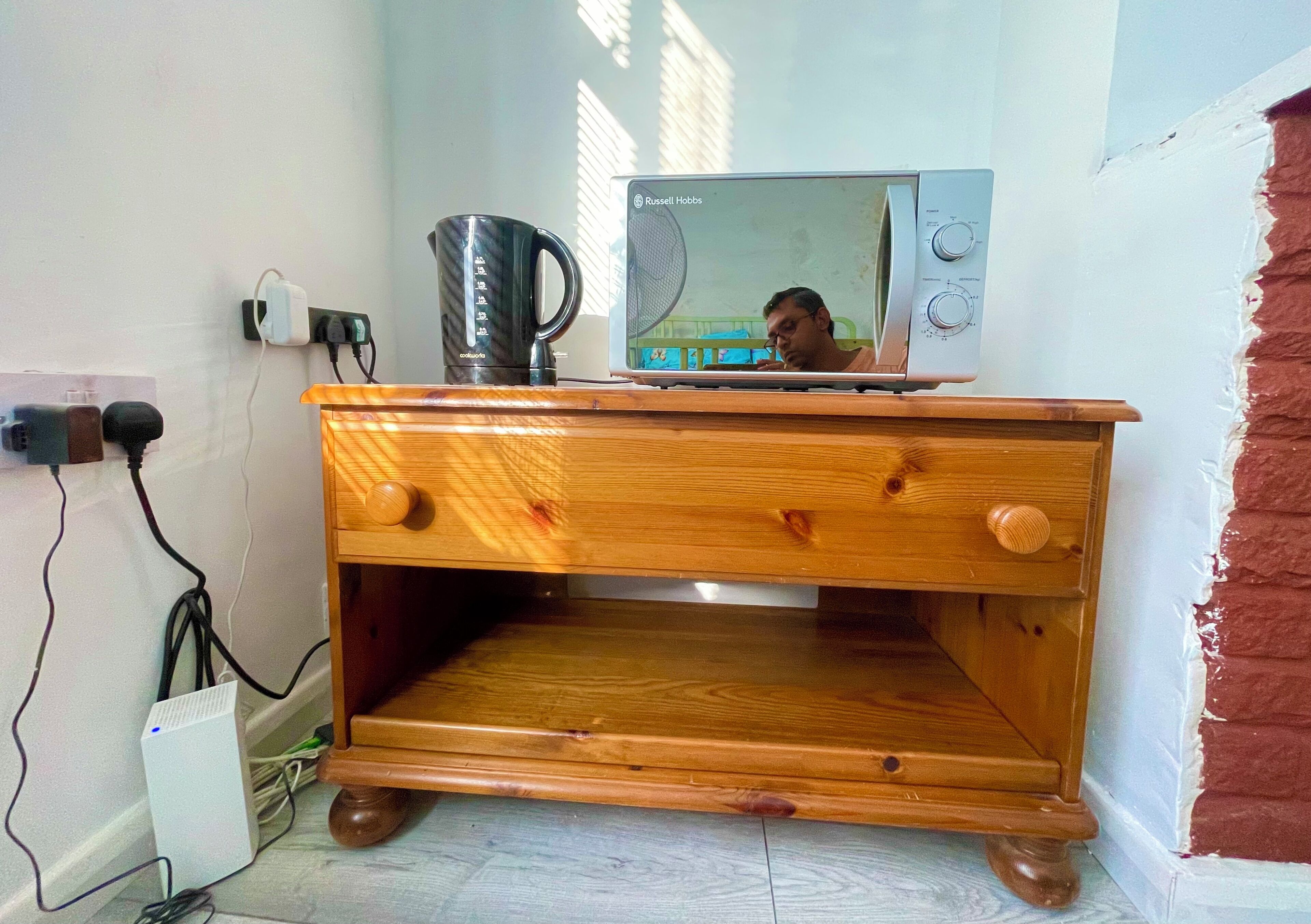 A wooden cabinet with a microwave on top, a kettle, and a power strip with cables.