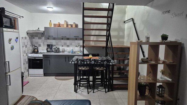 A compact kitchen with a stove, sink, and cabinets, a dining area with a table and chairs, a small sofa, and a wooden shelf with decorative items.