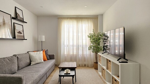 A modern living room with a grey sofa, a coffee table, a TV on a white stand, and a large window with sheer curtains. - Chic Escape Apartment (Godella)