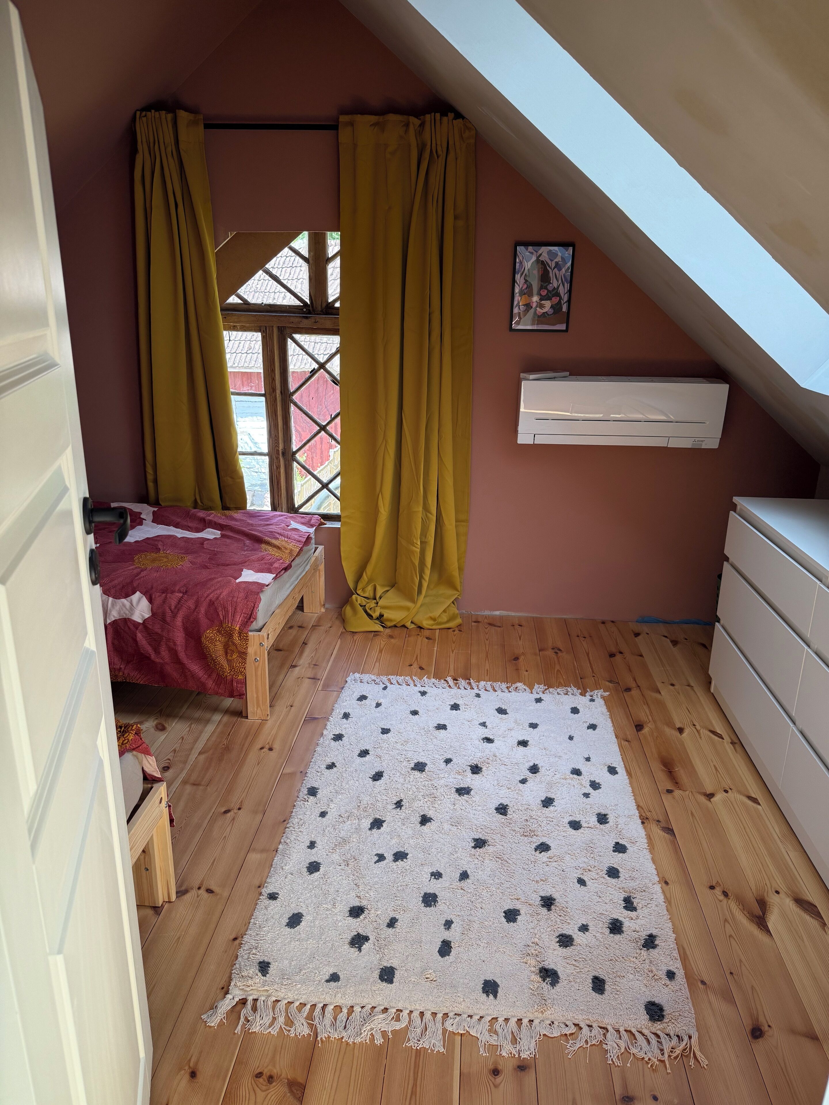 Een kleine, gezellige slaapkamer op zolder met een schuine kap, houten vloeren, een bed met een rood-geel gebeddeken, een witte airconditioner en een groot raam met gele gordijnen.