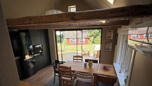 Een keuken met een houten tafel en stoelen, een inbouwoven en uitzicht op een rode woning door grote ramen.