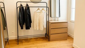 A room with a wooden floor, a clothing rack with jackets and a white shirt, a mirror, and a dresser.