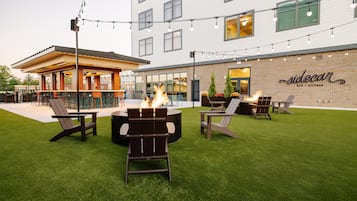 Un espacio al aire libre con fogones y sillones de descanso, un bar, y un edificio con fachada de ladrillo y un letrero que dice 'Siccar'.