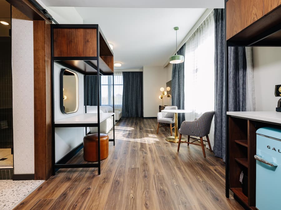 A hotel room with a wooden floor, a bed with a mirror, a desk, a chair, a coffee machine, and a blue cabinet.