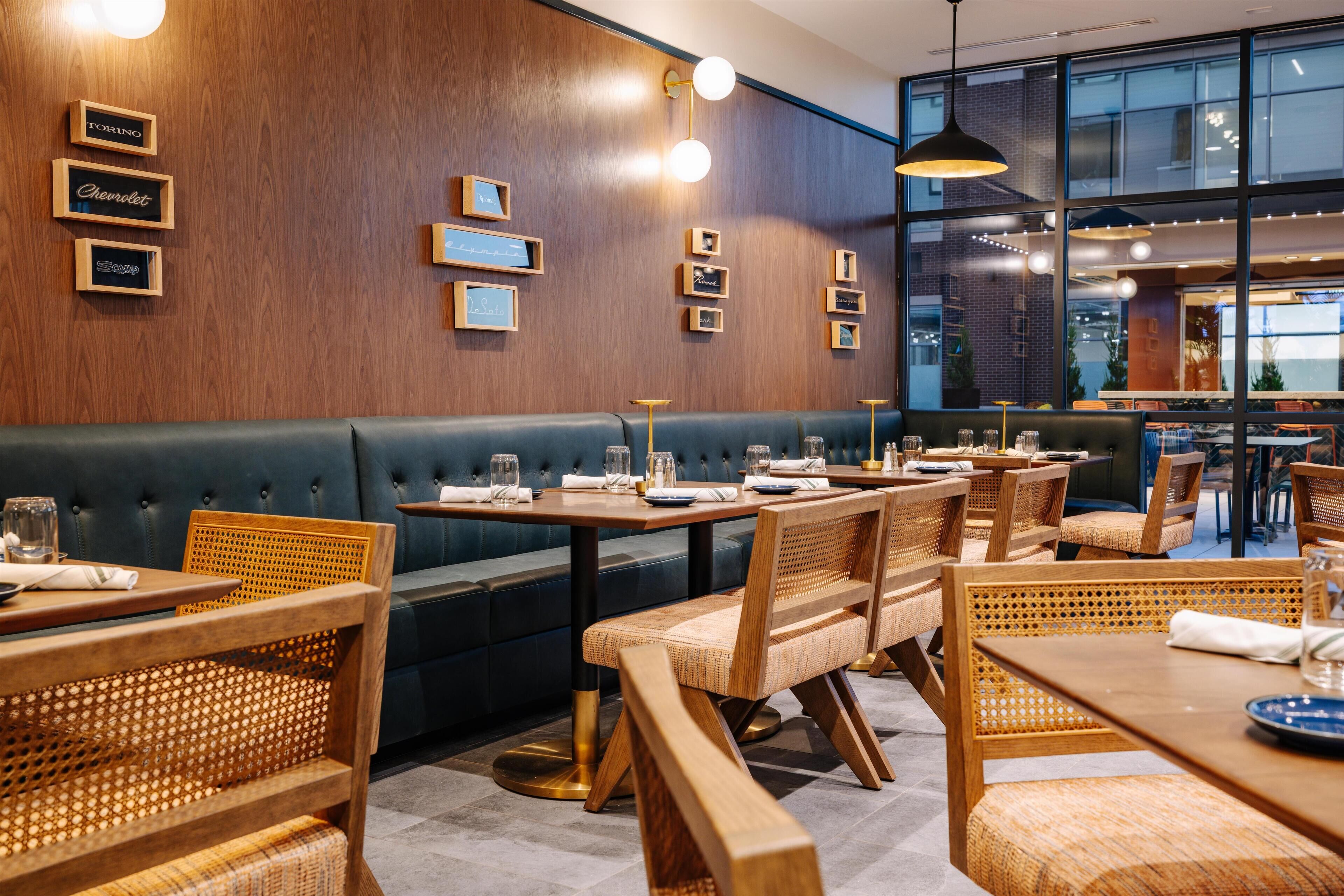 L’intérieur d’un restaurant présente des murs en bois, un coin salon avec des banquettes bleues et des tables accompagnées de chaises en osier.