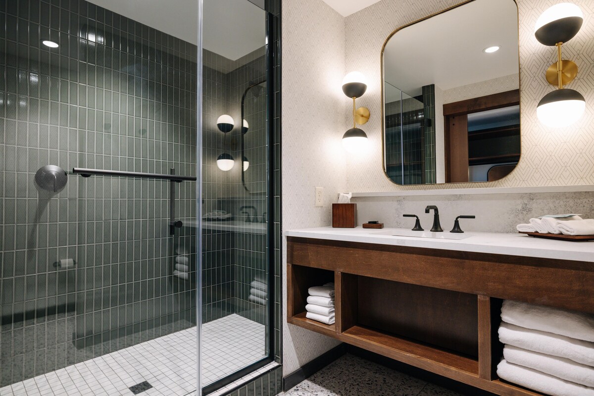 A modern bathroom with a glass shower enclosure, a wooden vanity, and a large mirror with wall-mounted lights.