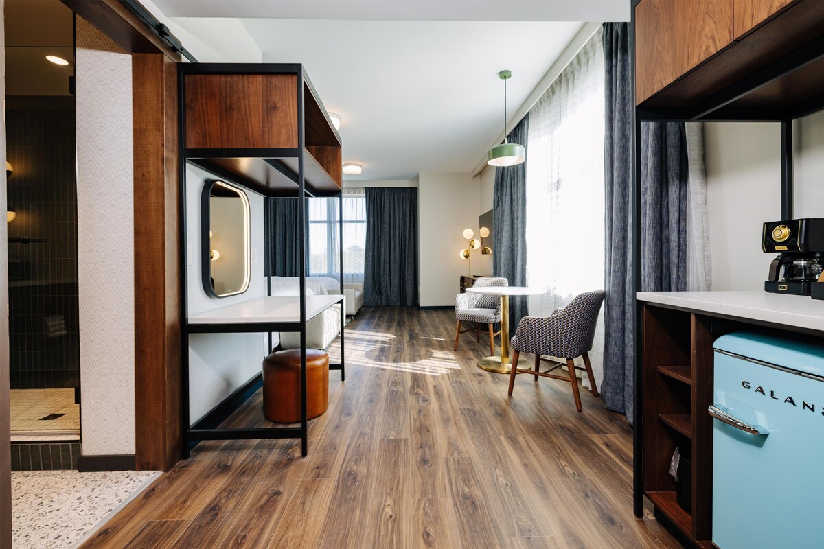 Quarto de hotel moderno com piso de madeira, cama, escrivaninha, cadeira e cozinha compacta.
