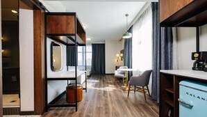 Habitació d'hotel moderna amb terra de fusta, un llit amb un mirall damunt, una taula petita amb cadires, un refrigerador blau i una finestra gran amb cortines.