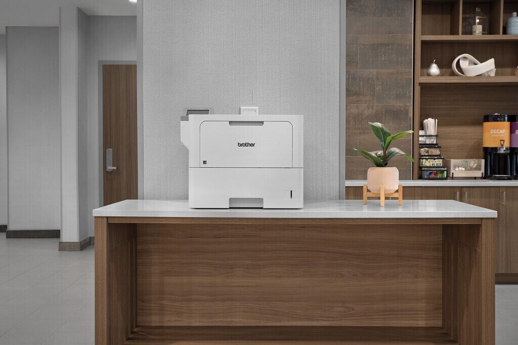 A Brother printer on a white countertop in a modern kitchen with wooden cabinets and a potted plant.