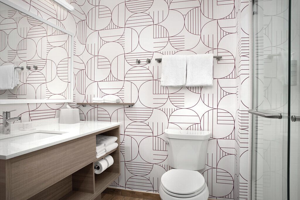 Un baño moderno con pared de diseño geométrico, un lavabo blanco, un inodoro y una cabina de ducha con mampara de cristal.