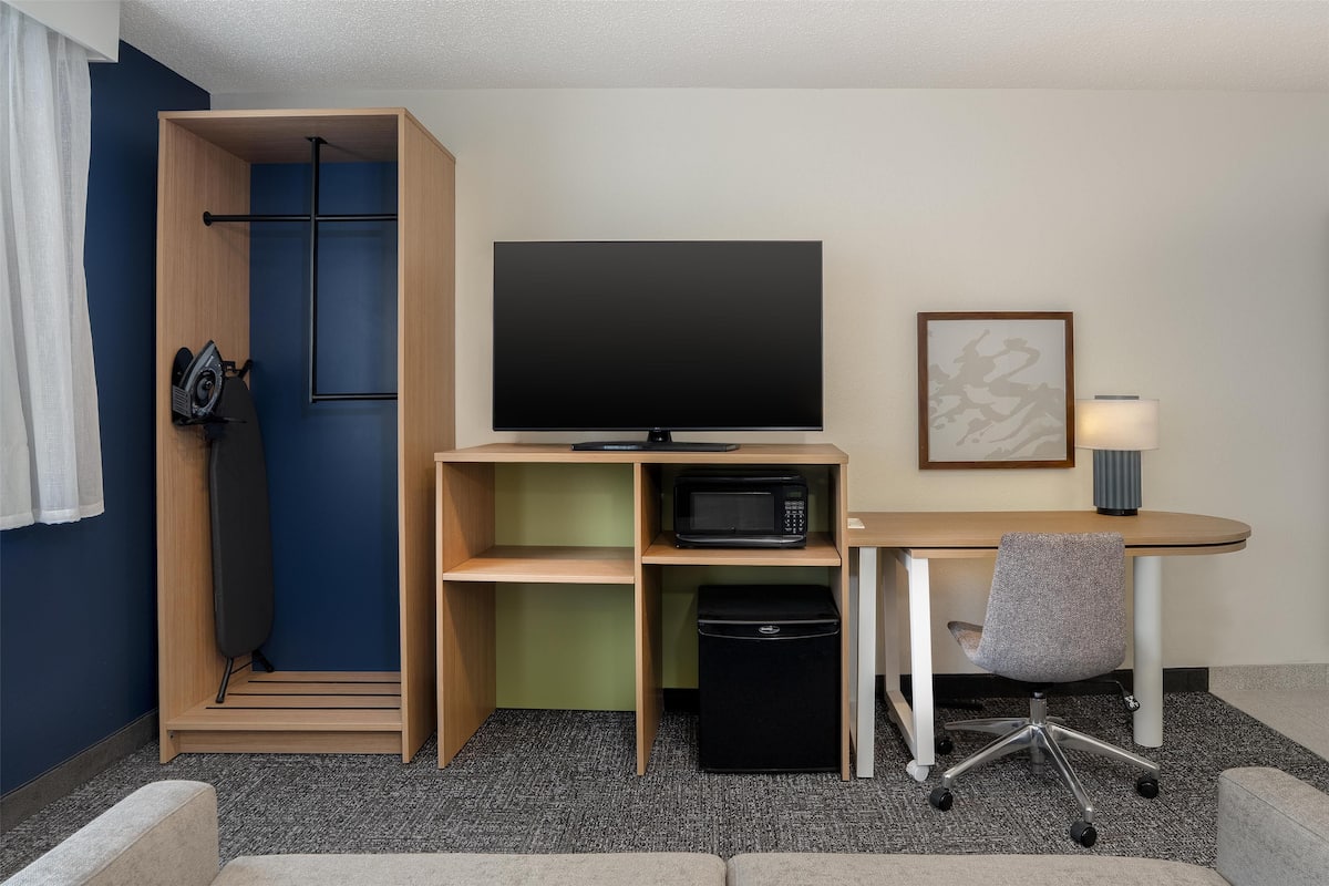Une chambre d’hôtel compacte comprenant une armoire en bois, un téléviseur à écran plat, un four à micro-ondes, une petite poubelle, un bureau avec une chaise et une lampe.