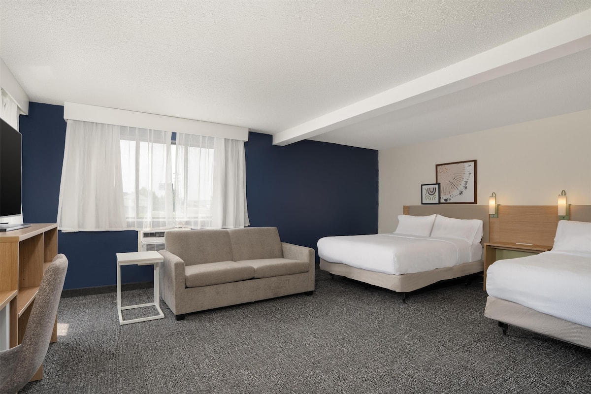 Une chambre d’hôtel avec un canapé, deux lits simples, une télévision, une petite table et une fenêtre avec des rideaux.