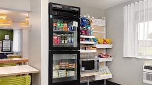 A kitchenette with a refrigerator stocked with beverages, a microwave, and a small refrigerator with packaged food. There are shelves with snacks and a window with curtains. - Spark By Hilton Carlisle South (Carlisle)