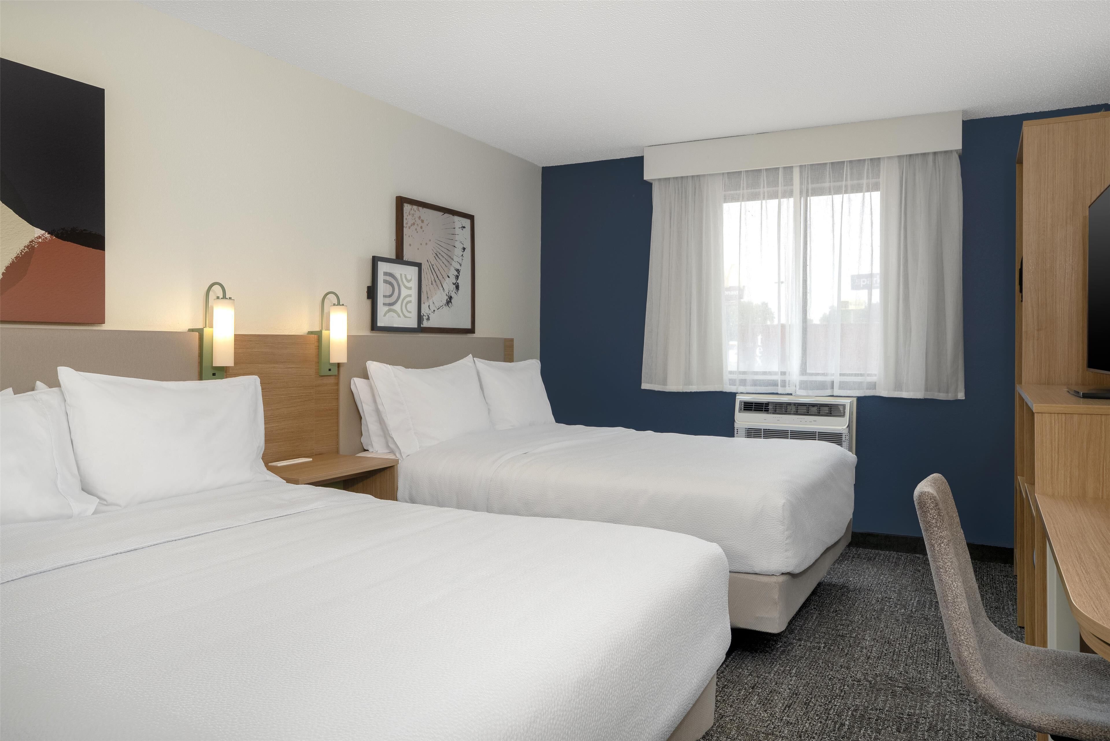 Ein Hotelzimmer mit zwei Betten, einem Schreibtisch, einem Stuhl, einem Fenster mit Vorhängen und Wandbildern.