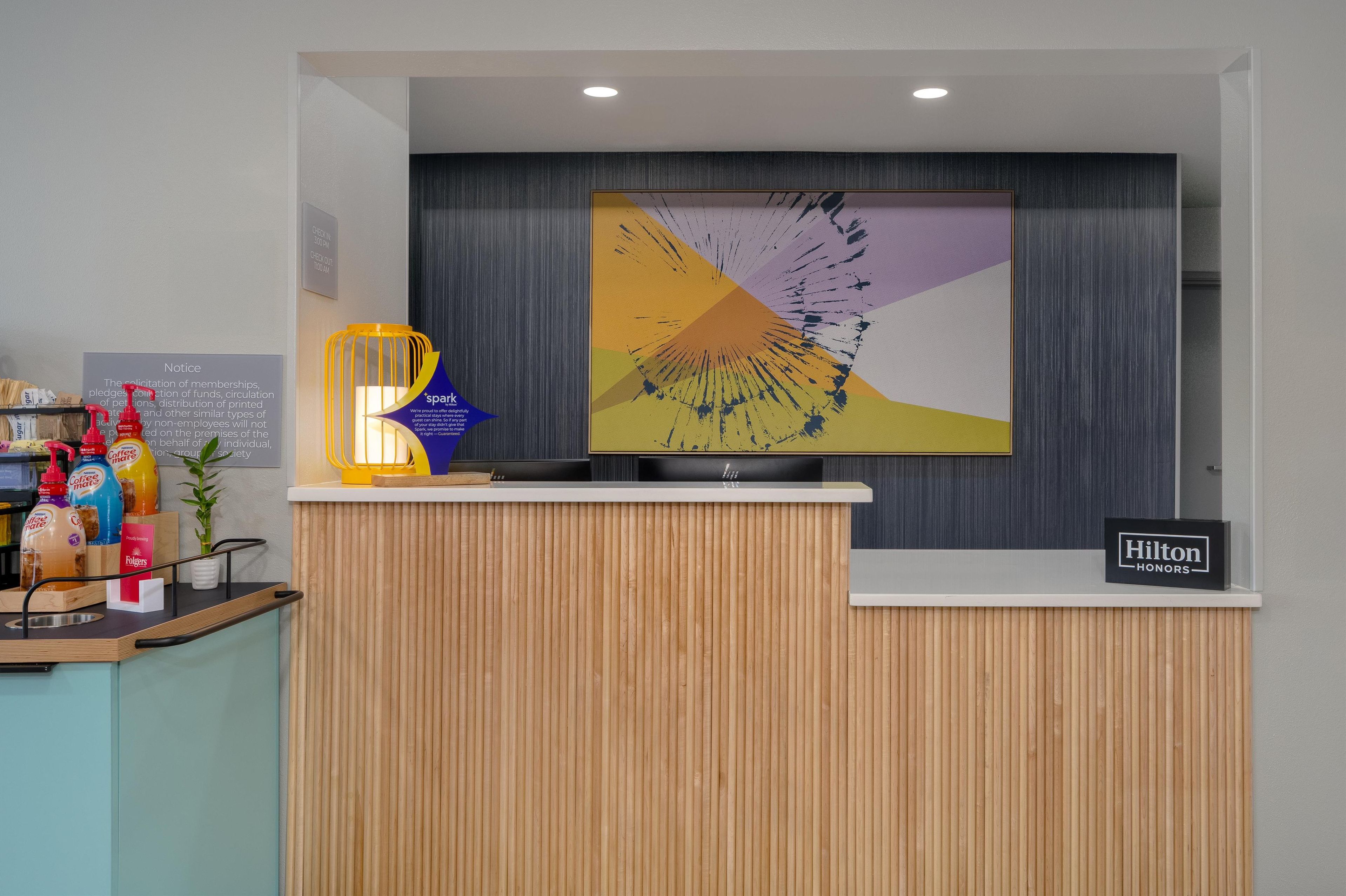 A hotel reception desk with a modern design, a large abstract artwork above it, and a sign indicating it's a Hilton Honors hotel.