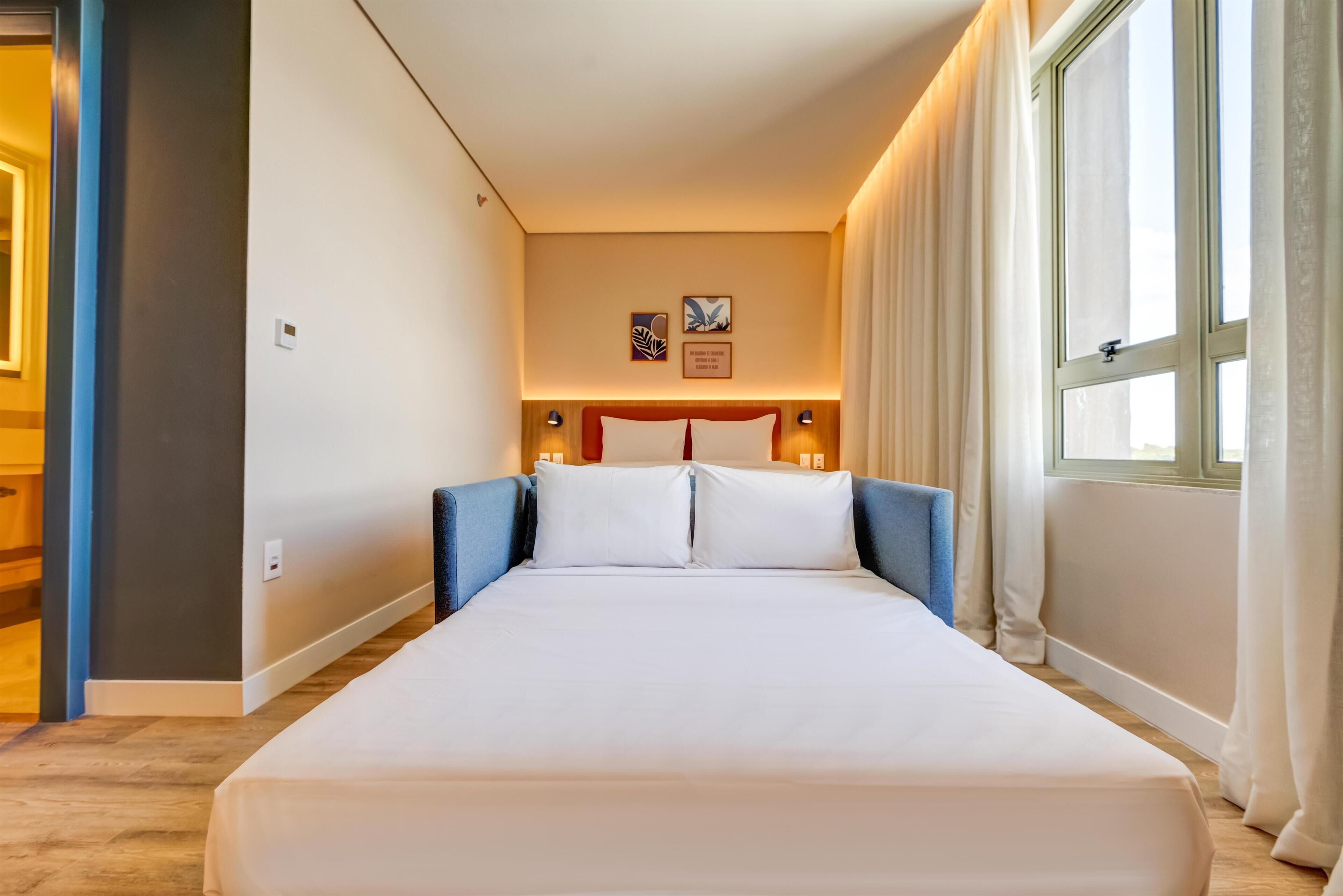A hotel room with a large bed, a headboard with a red accent, two wall-mounted lamps, and a window with curtains.