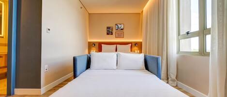 Een hotelkamer met een groot bed, een hoofdbord met rode accenten, twee wandlampen en een raam met gordijnen.