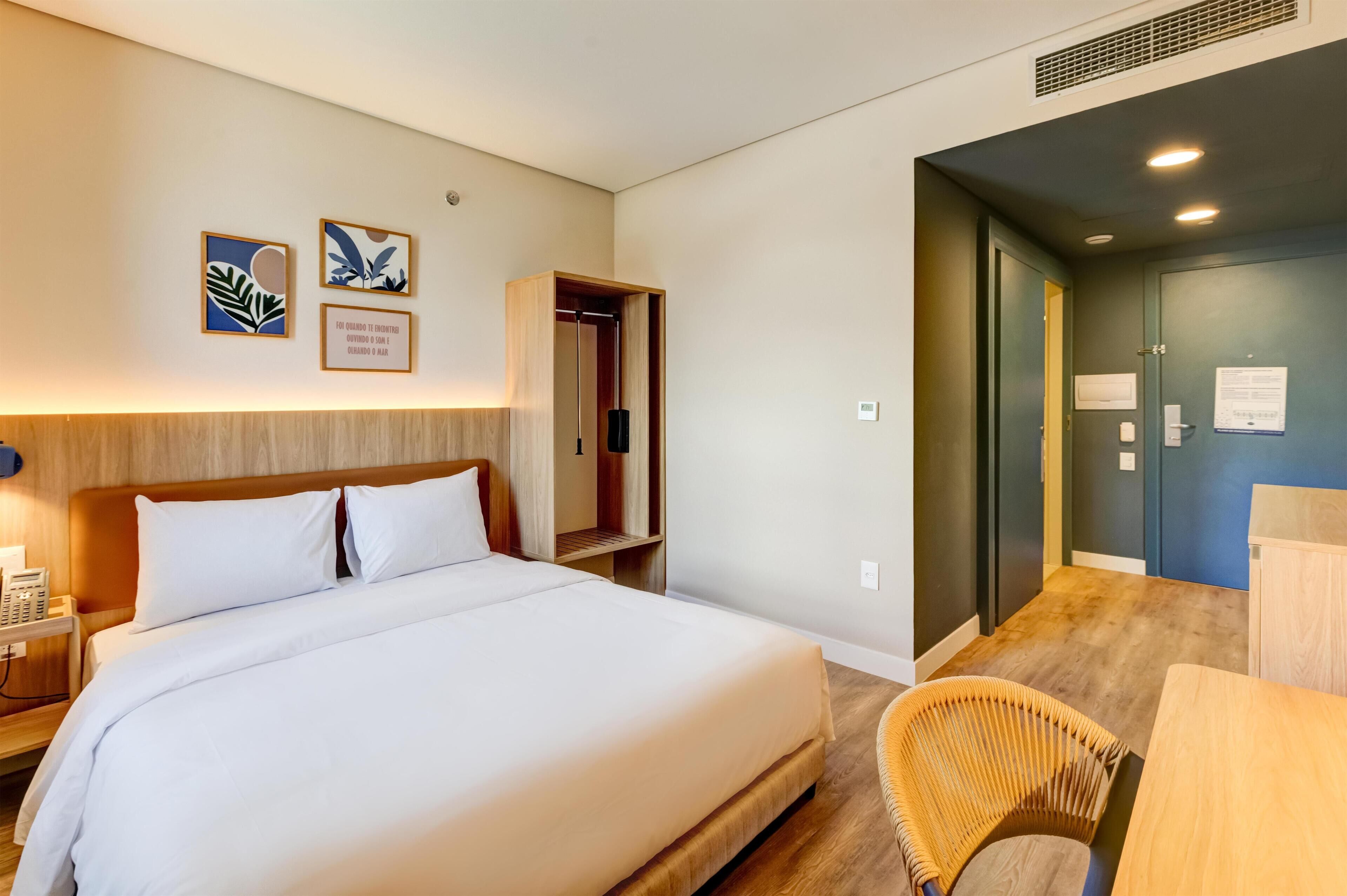 Quarto de hotel com uma cama grande, telefone ao lado da cama, cabeceira de madeira, guarda-roupa, escrivaninha e uma cadeira.