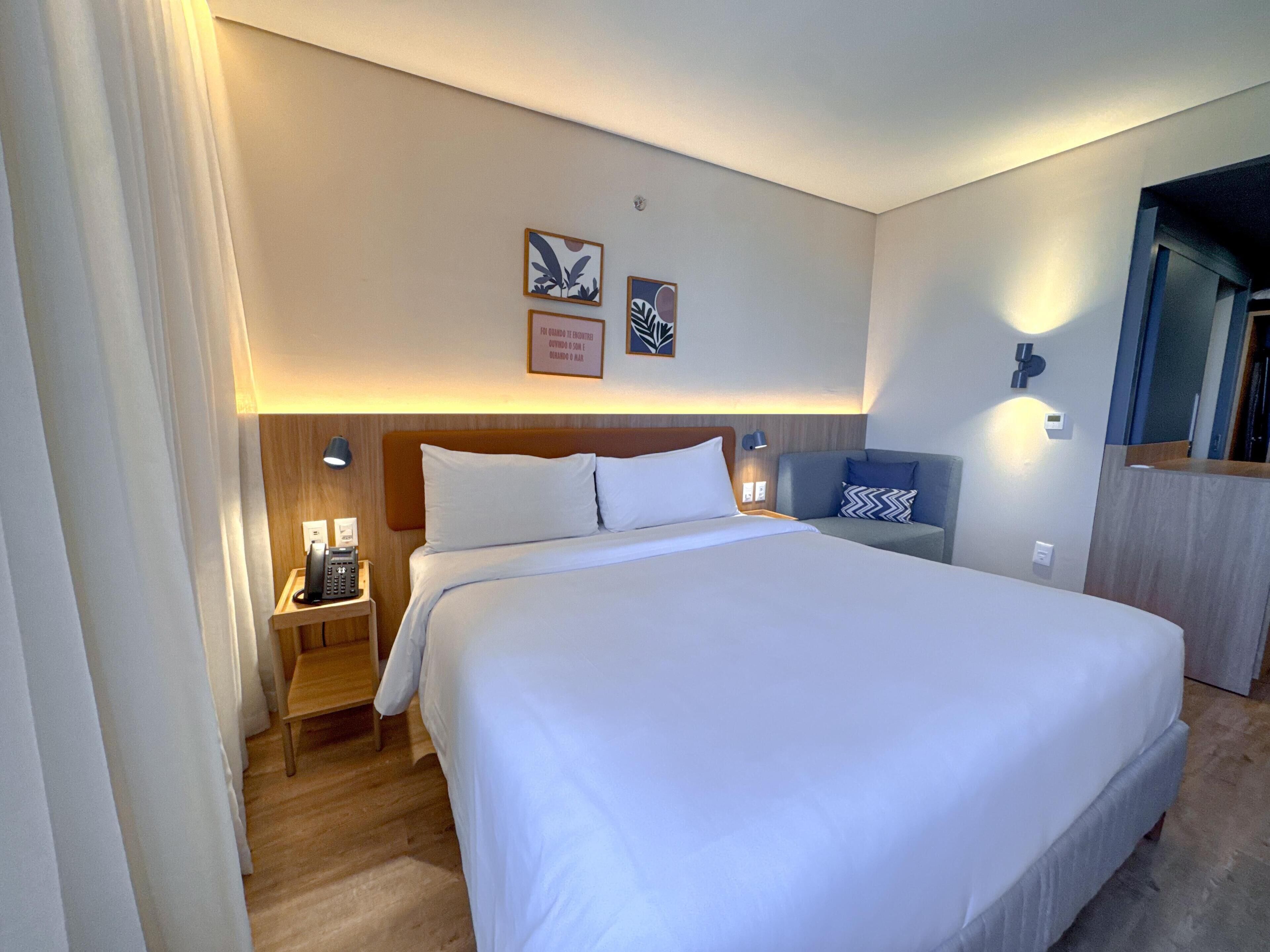 A hotel room with a large bed, bedside tables, a nightstand, a sofa, and wall-mounted lights.