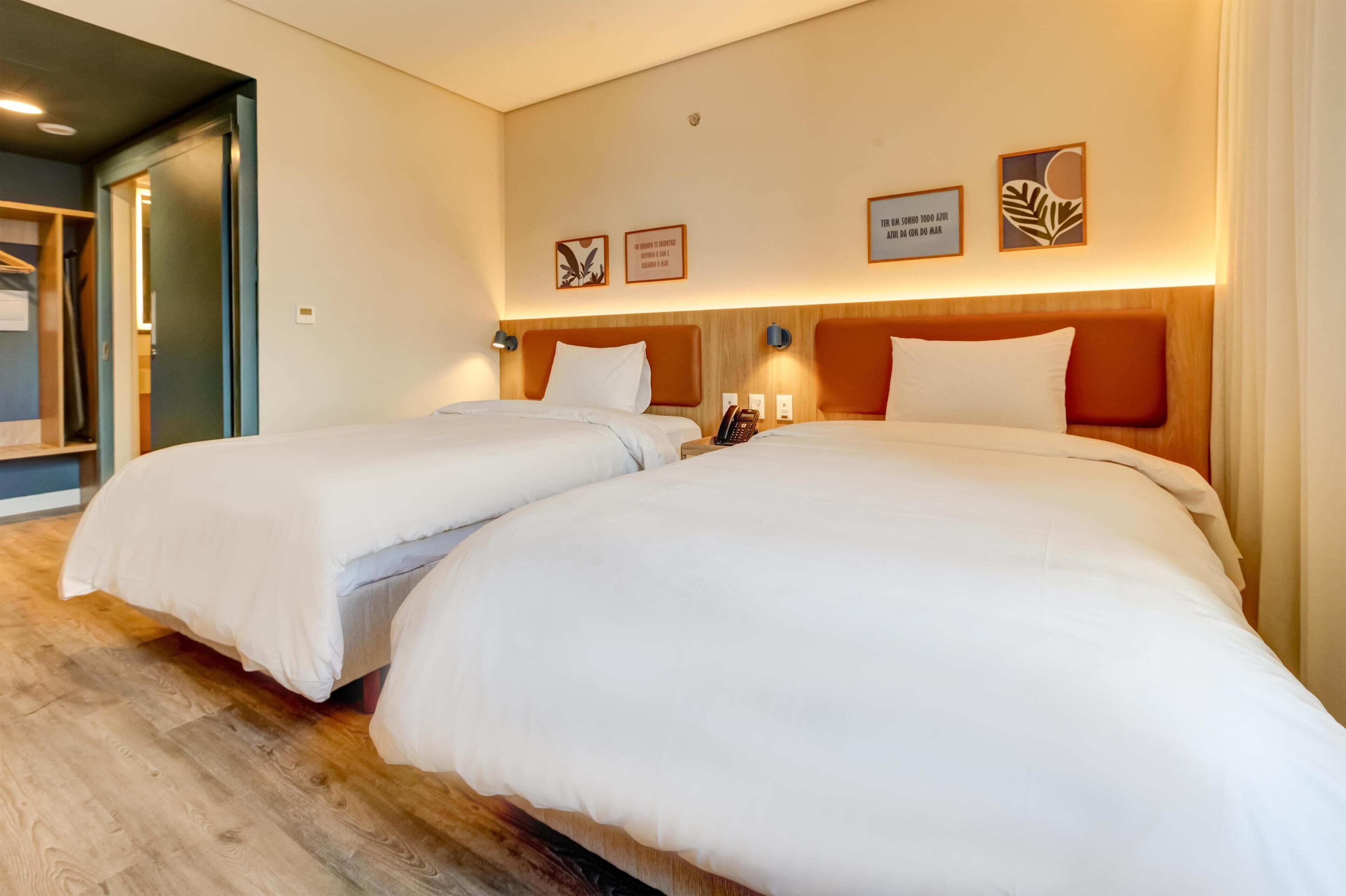 Quarto de hotel com duas camas, cabeceira de madeira, luminárias de cabeceira e quadros nas paredes.