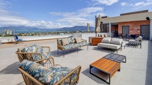 A rooftop terrace with wicker furniture, a wooden coffee table, and a view of the city and mountains. - Hampton By Hilton Caraguatatuba Serramar (Caraguatatuba)