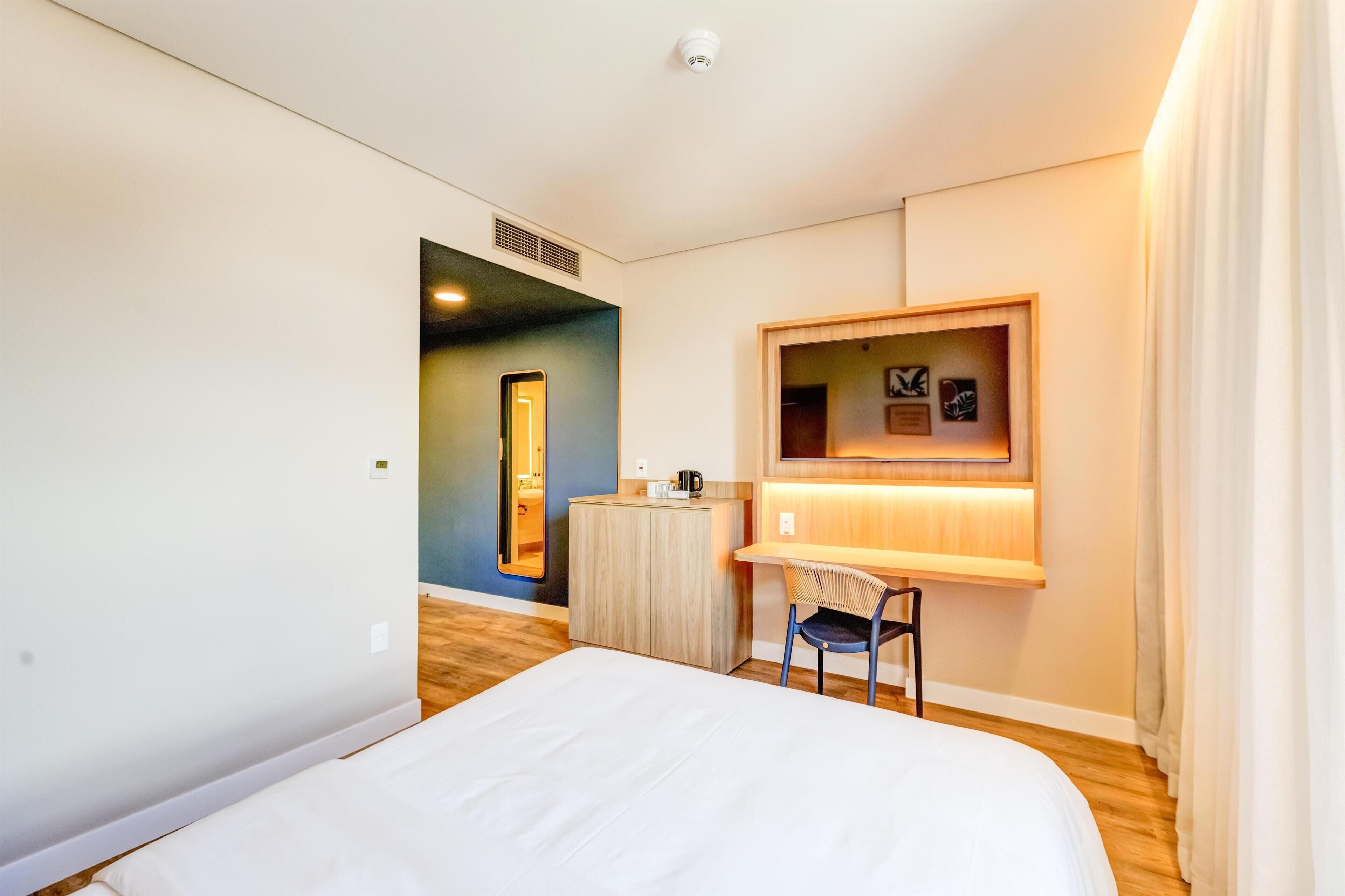 Ett hotellrum med en säng, ett skrivbord med en stol, ett litet bord, en spegel och en vägghylla med bilder.