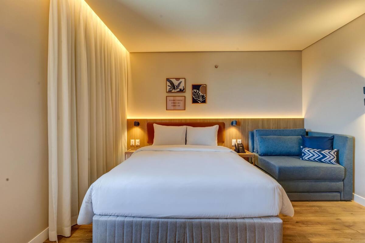Quarto de hotel com uma cama grande, uma mesa de cabeceira, um criado-mudo, um sofá azul e duas imagens emolduradas na parede.