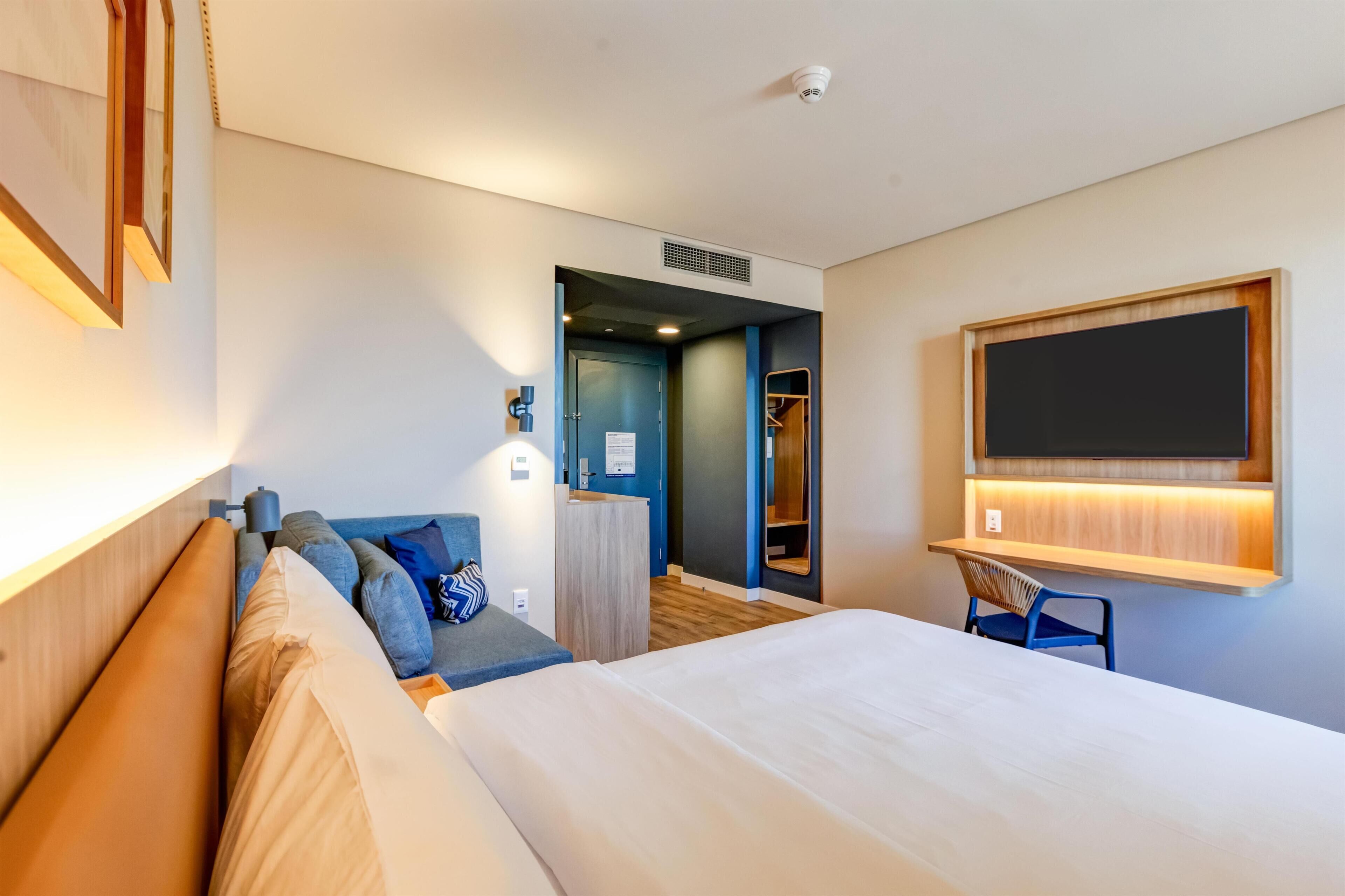 Habitación de hotel con una cama grande, un escritorio con una silla, un armario y un baño visible a través de una puerta abierta.