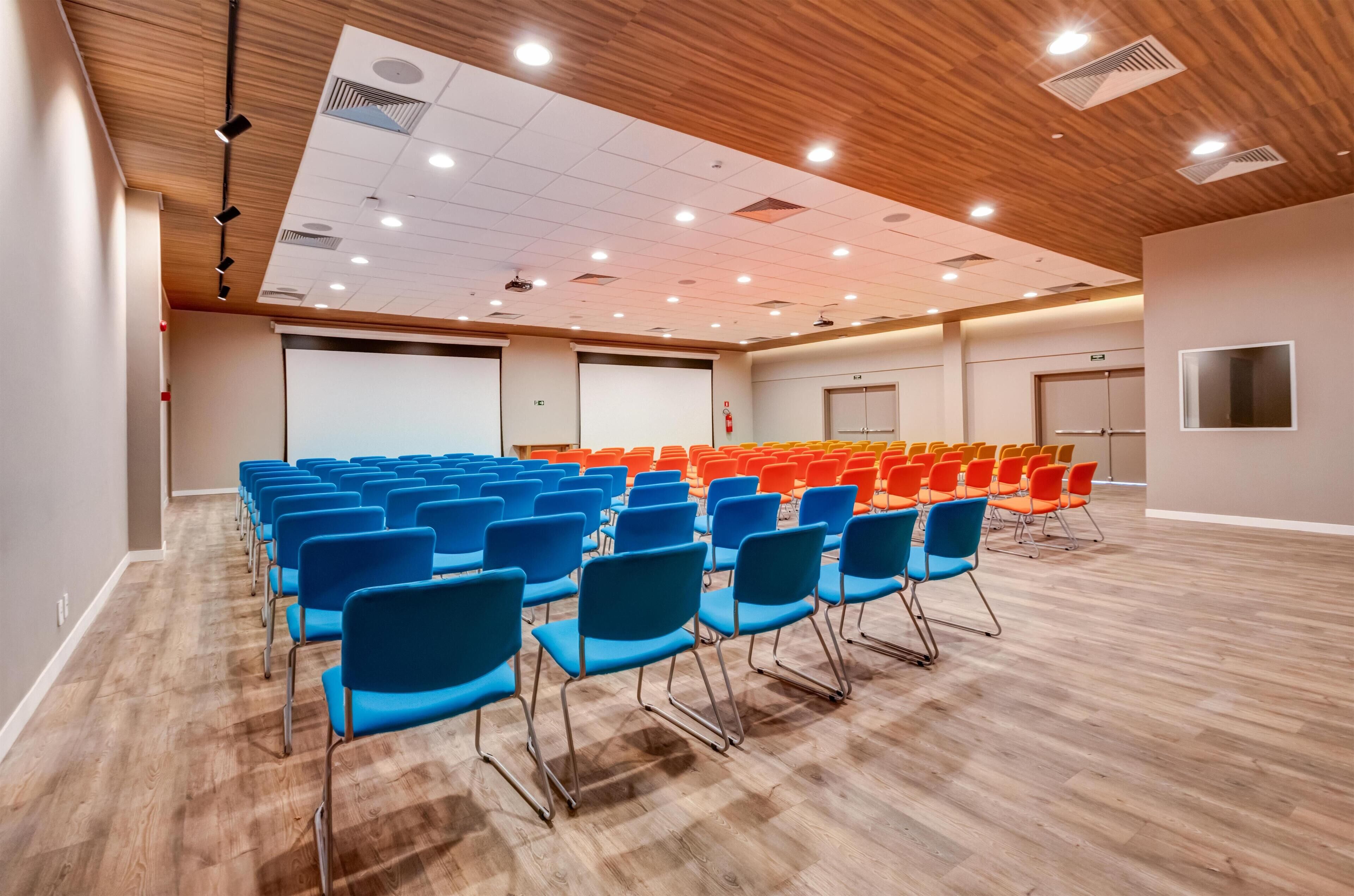 Ein Konferenzraum mit Reihen von blau und orangefarbenen Stühlen, Holzböden und einer weißen Leinwand im Vordergrund.
