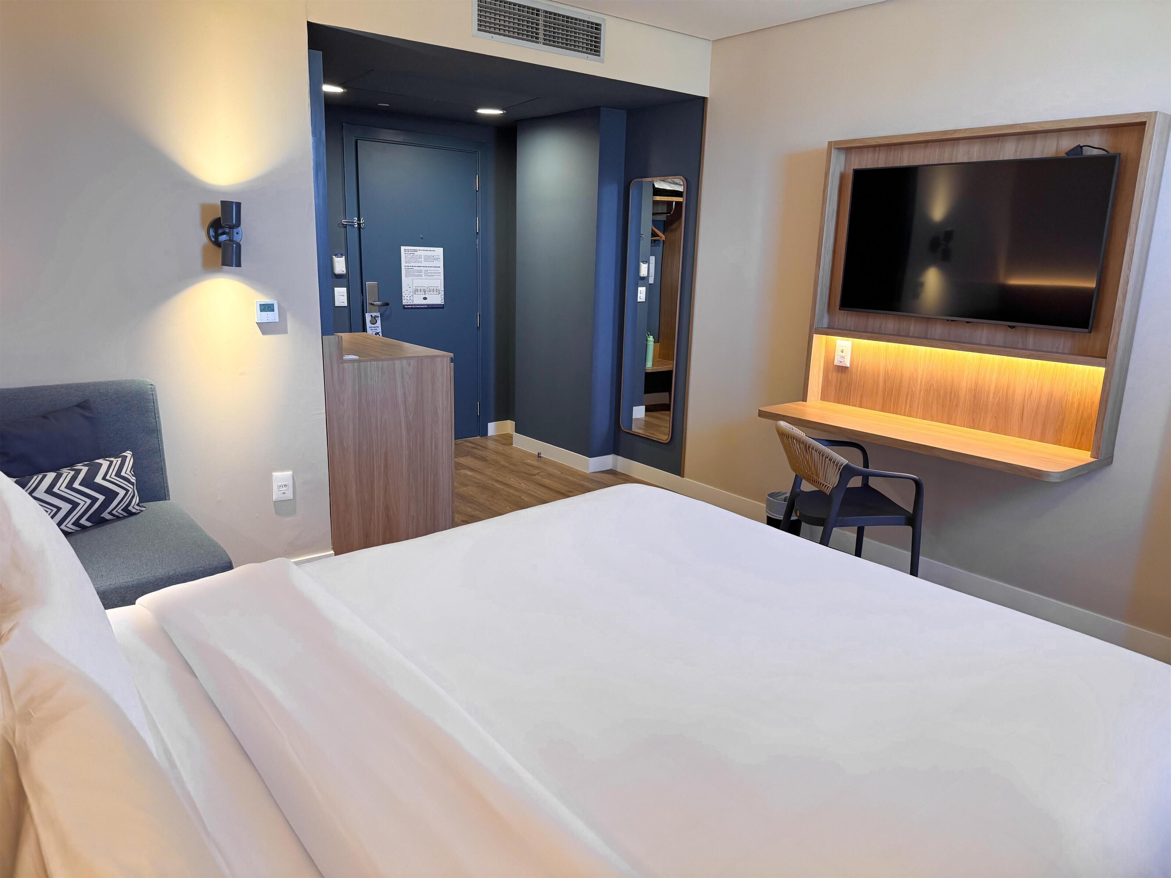 A hotel room with a bed, a desk with a chair, a TV, a wardrobe, and a mirror.