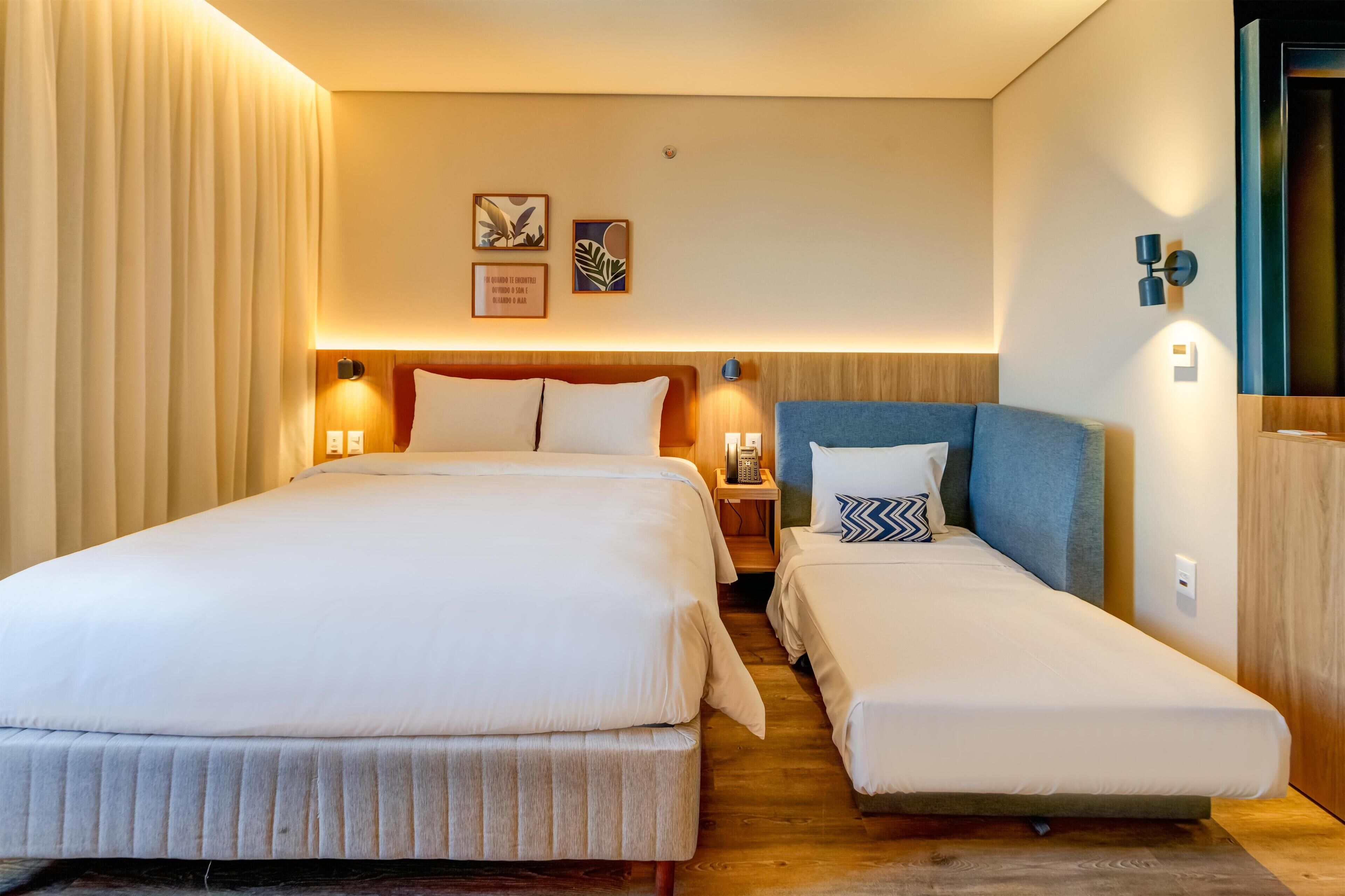 A hotel room with a large bed, a smaller bed, a nightstand, a lamp, and wall art.