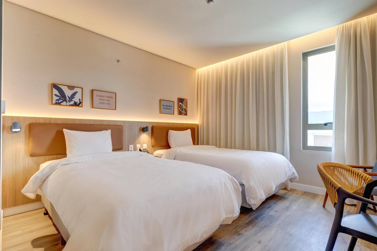 Quarto de hotel com duas camas, uma cabeceira de madeira, uma janela com cortinas e uma cadeira de vime.
