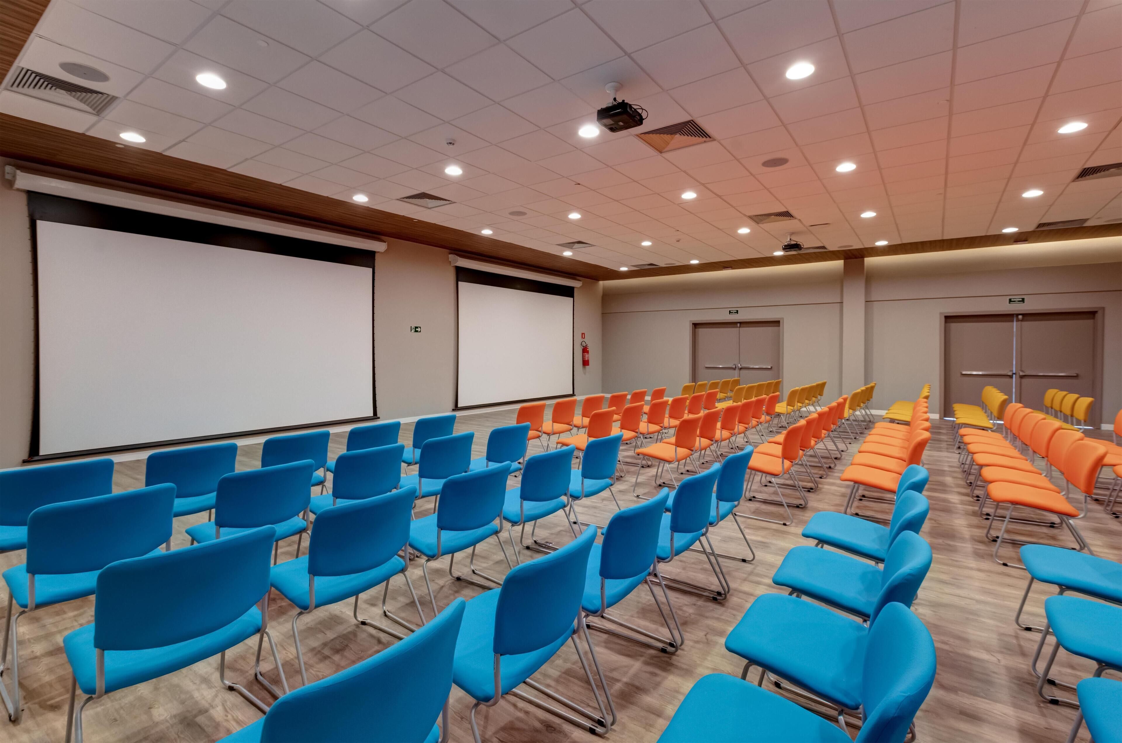 Ein Konferenzraum mit Reihen von blau und orangefarbenen Stühlen, einer weißen Leinwand und einer Holzzahl.
