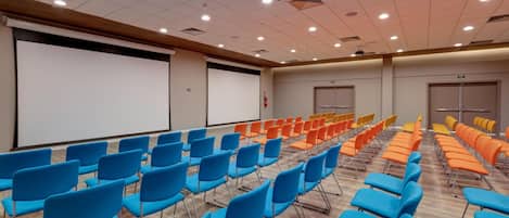 A conference room with rows of blue and orange chairs, a white screen, and a wooden ceiling.