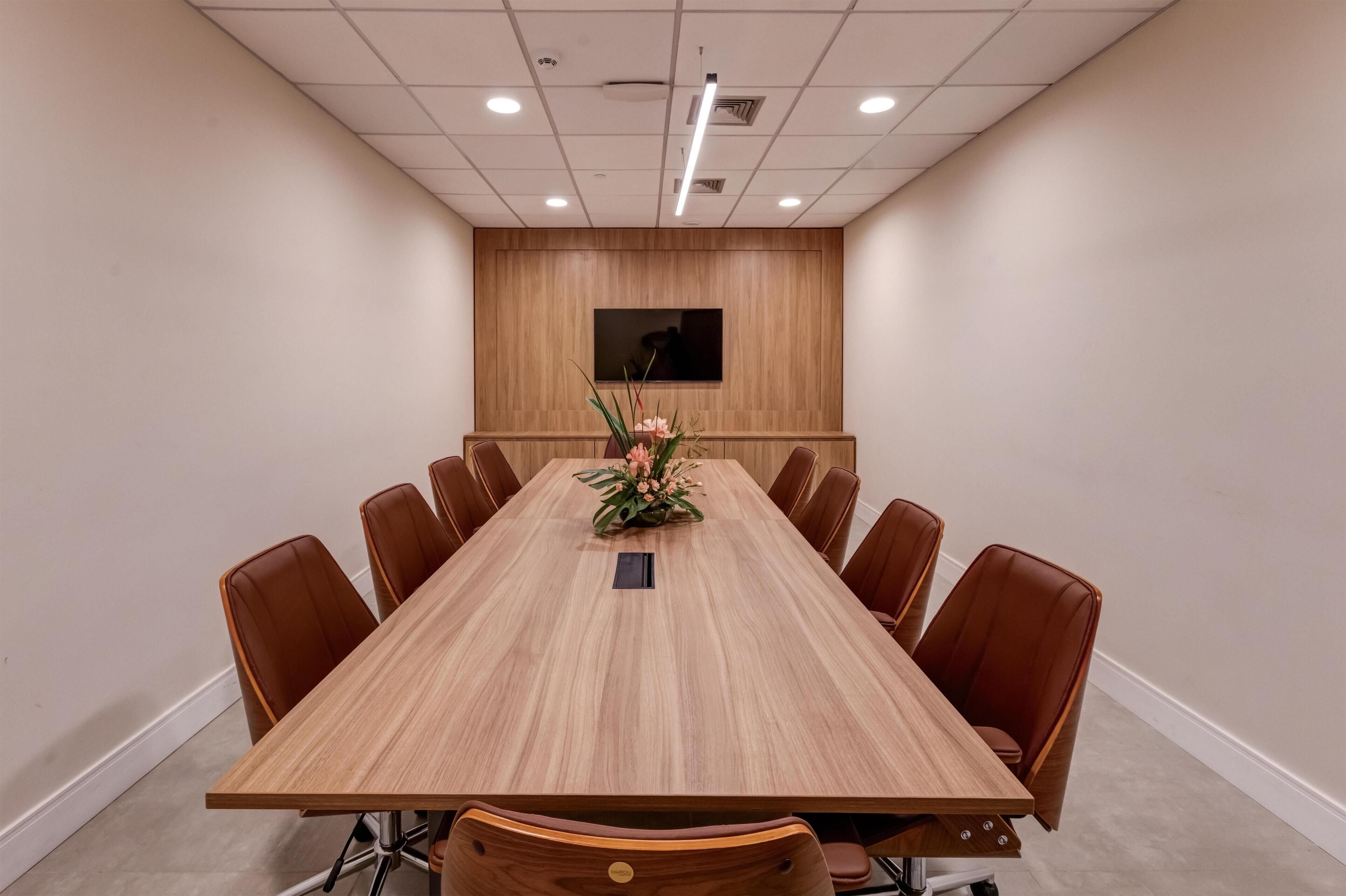 A conference room with a long wooden table, brown leather chairs, a flower centerpiece, and a mounted screen on the wall.