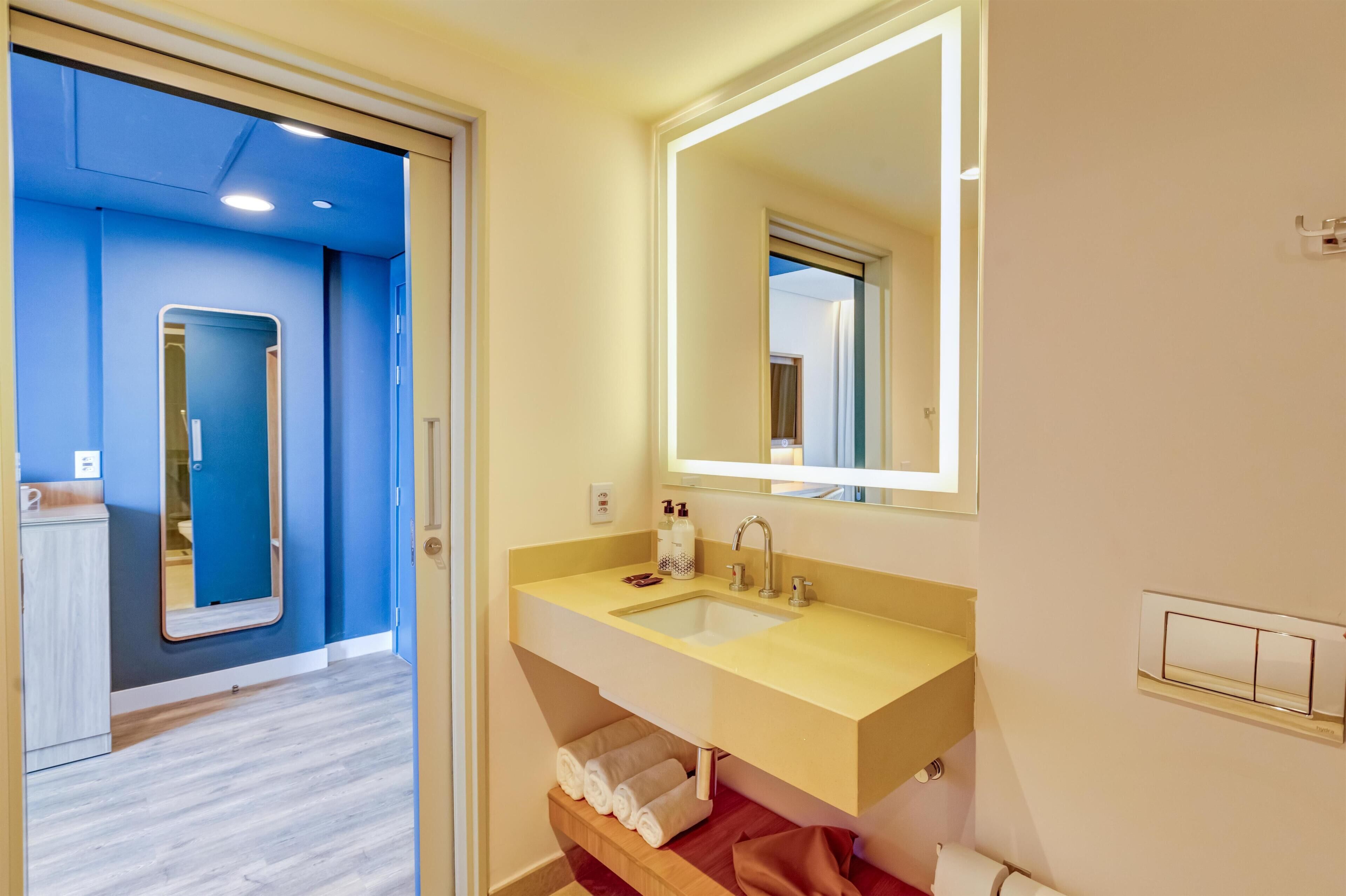 A modern bathroom with a yellow sink, a large mirror, and a wooden shelf with towels.