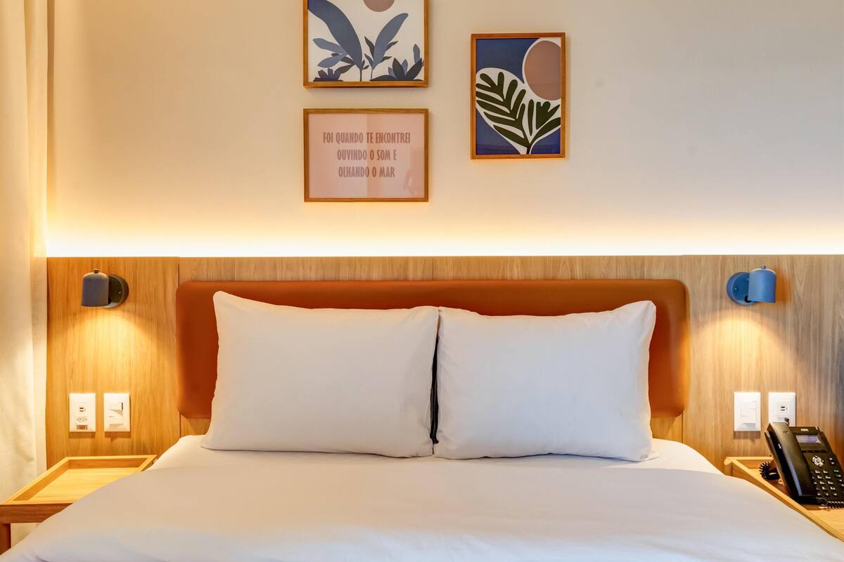 Quarto de hotel com cabeceira de madeira, duas almofadas brancas, mesa de cabeceira com telefone e duas luminárias fixadas na parede.