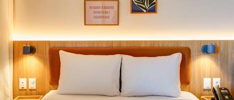 Habitación de hotel con cabecera de madera, dos almohadas blancas, una mesita de noche con teléfono y dos lámparas fijadas a la pared.