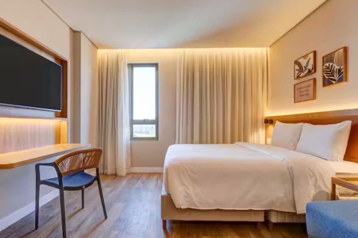 A hotel room with a bed, a desk, a chair, a TV, and a window with curtains.