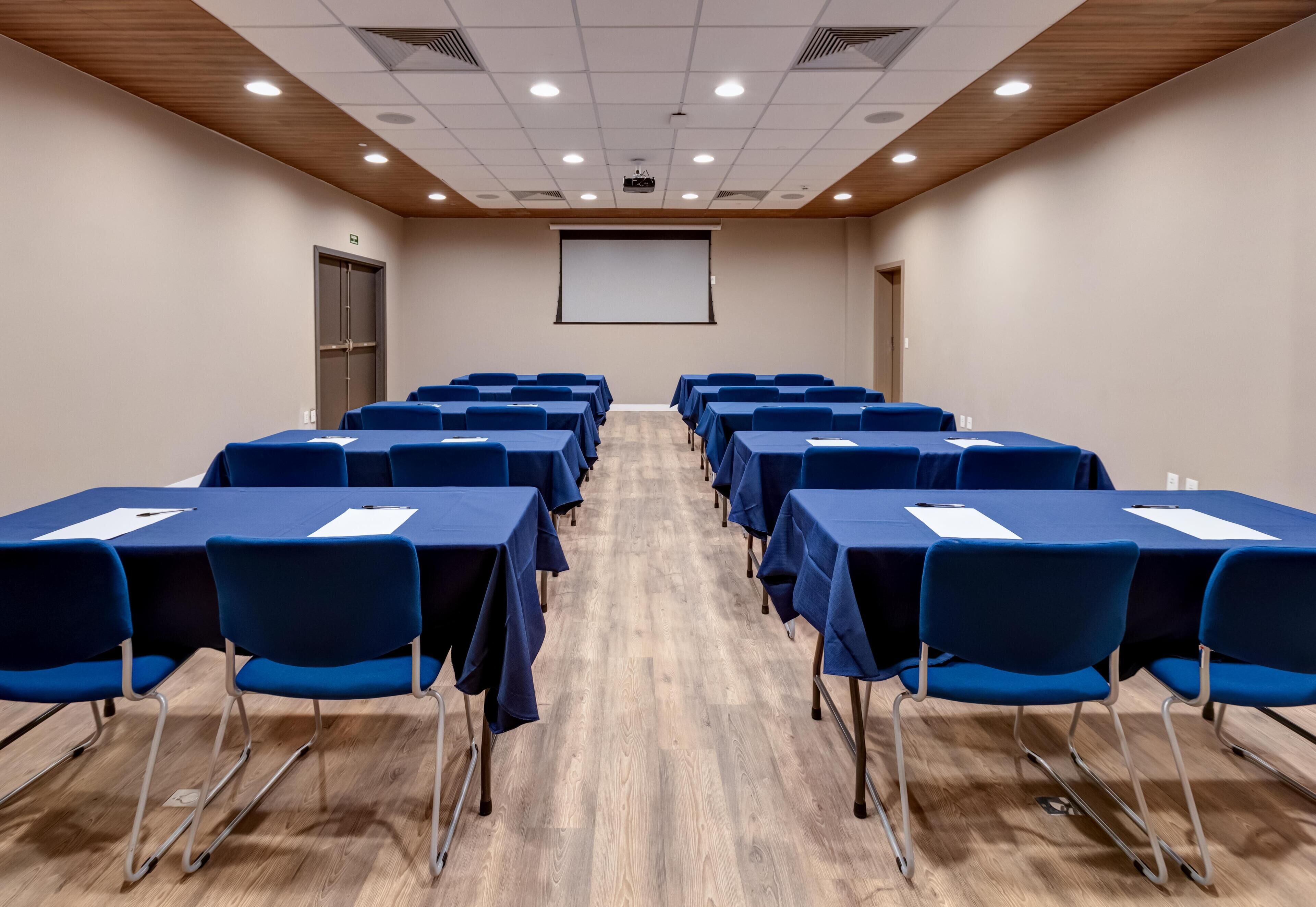 A conference room with rows of blue tables and chairs, a projector screen, and a wooden ceiling.