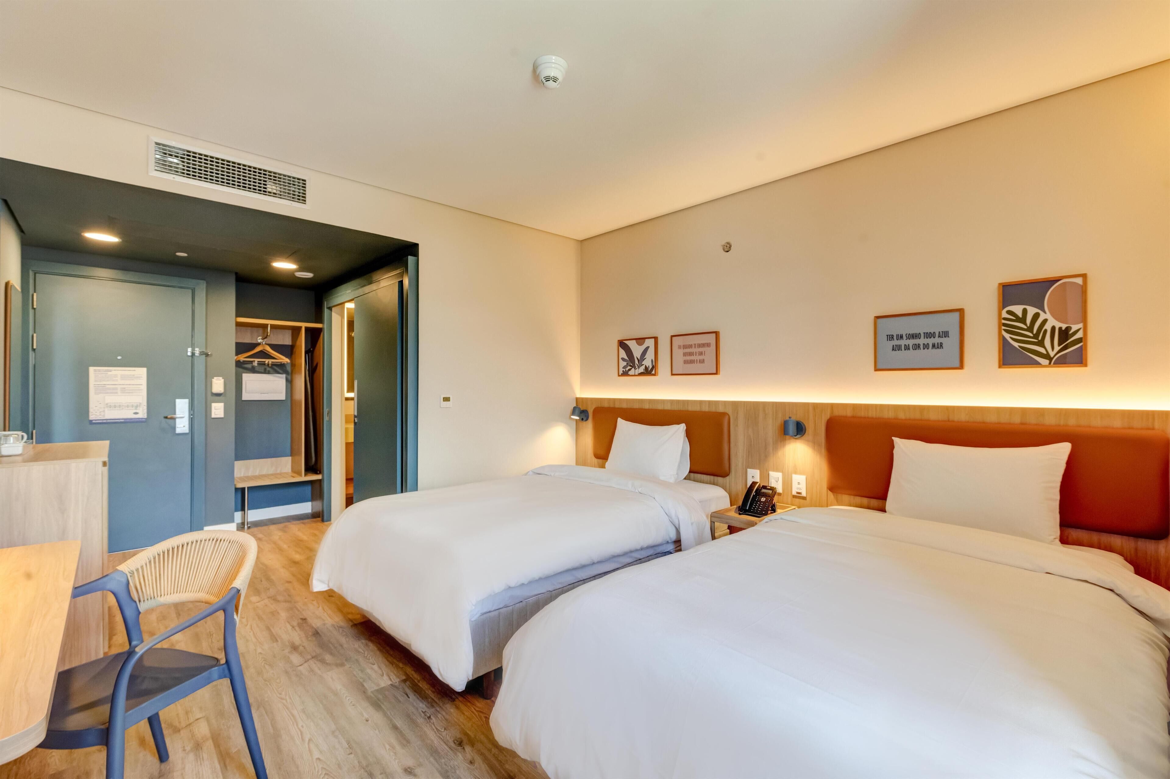 Habitación de hotel con dos camas, un escritorio y una silla. Se observa un armario y un espejo en una pared.