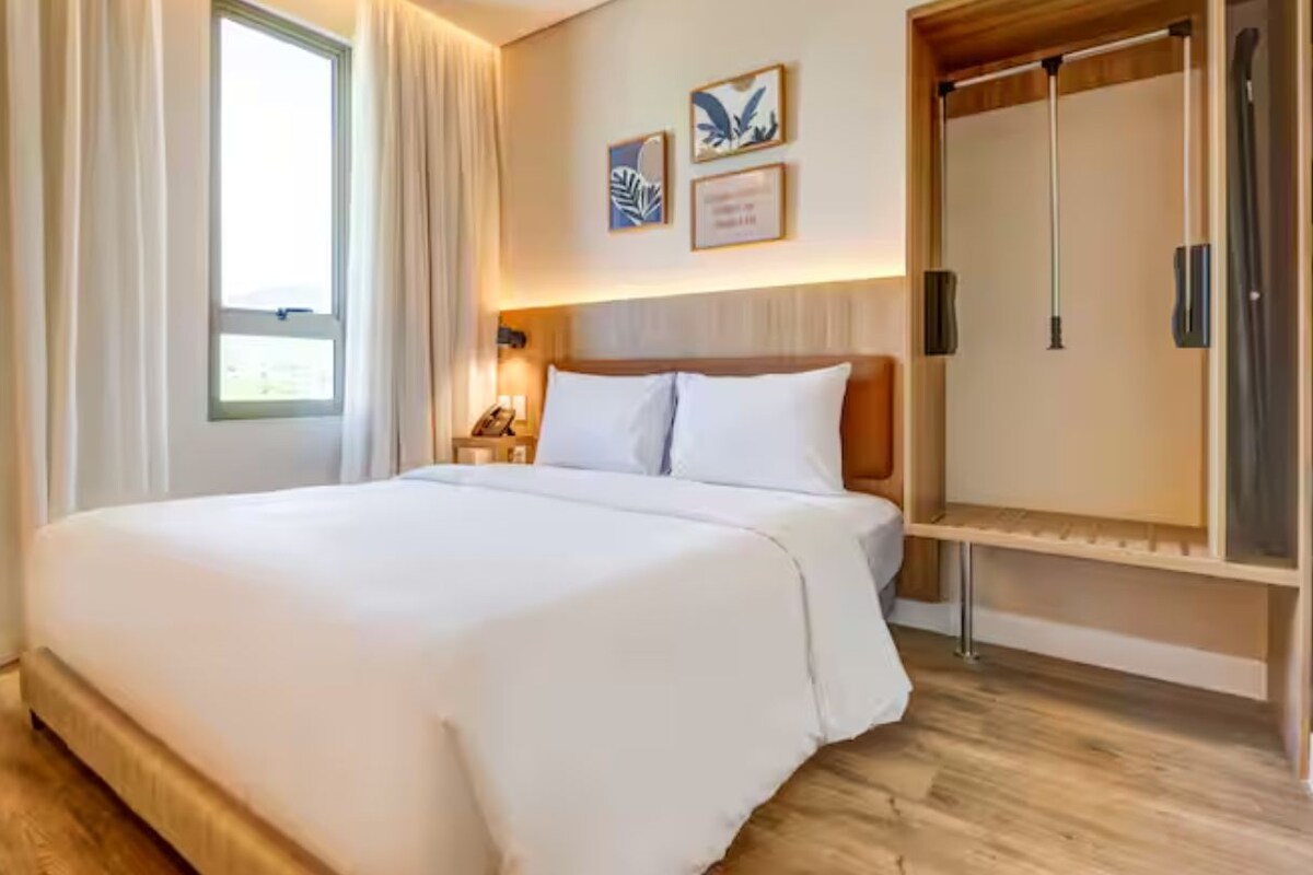 A hotel room with a large bed, a wardrobe, a bedside table with a lamp, and a window with curtains.