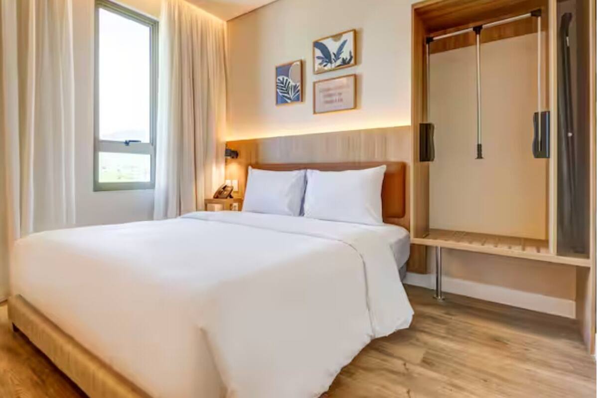 Quarto de hotel com uma cama grande, um guarda-roupa, uma mesa de cabeceira com abajur e uma janela com cortinas.