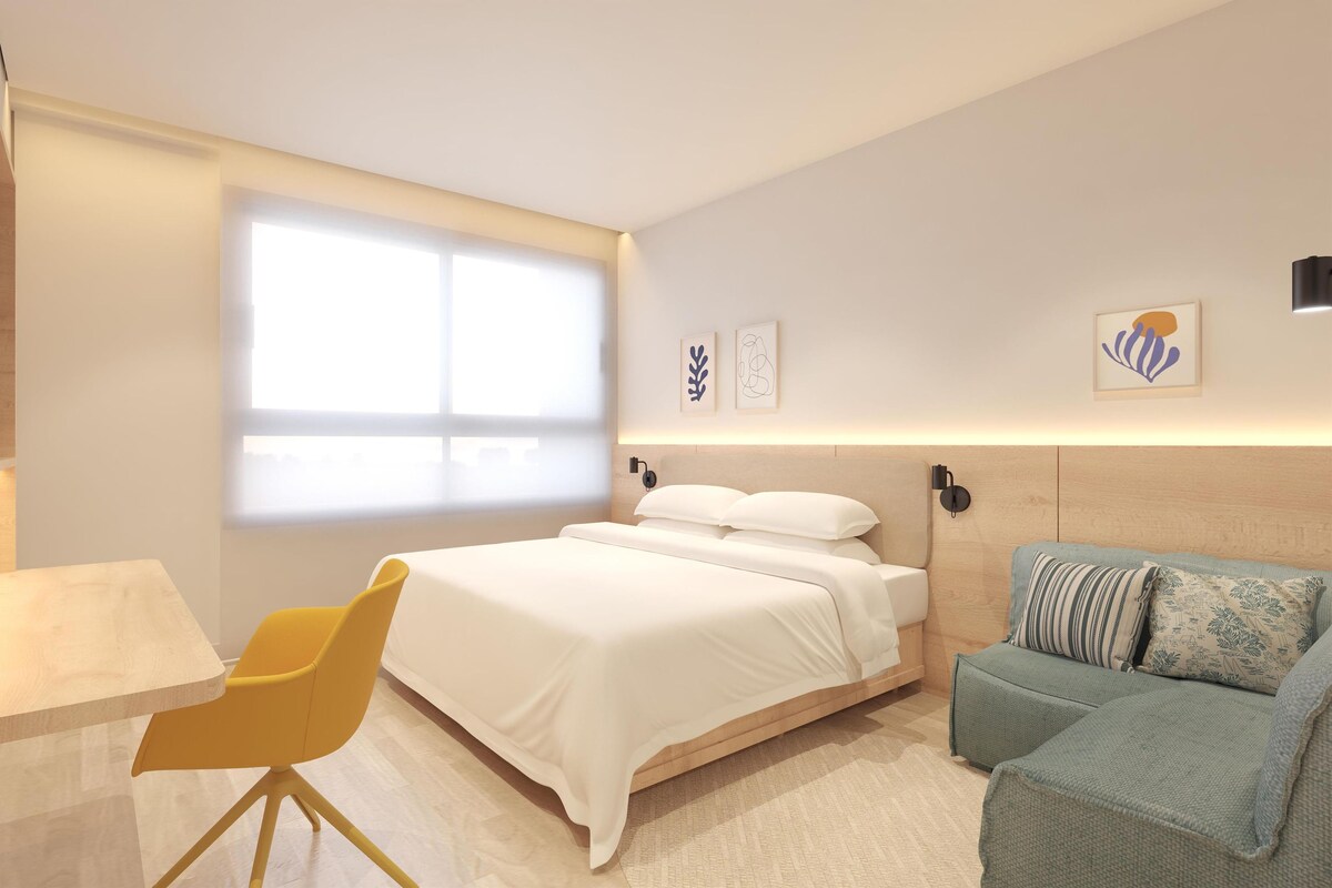 A modern hotel room with a bed, a desk, a chair, and a sofa.