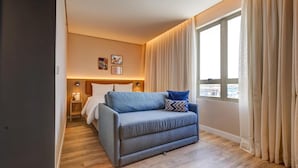 A modern hotel room with a grey sofa, a bed with white bedding, a nightstand with a lamp, and a large window with curtains. - Hampton By Hilton Caraguatatuba Serramar (Caraguatatuba)